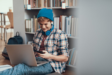 Positive student sitting with his laptop and looking at smartphone