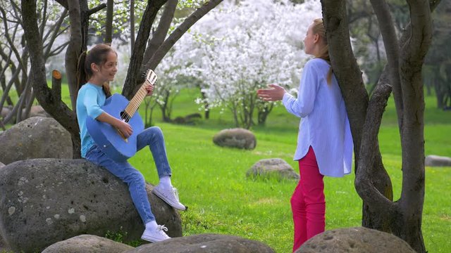 two cheerful little girlfriends playing music on guitar spring in the Park. beautiful idyllic scene in nature