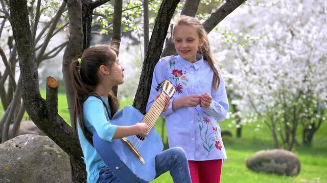 Happy and positive two school girlfriend playing music and singing in the nature Park. one young girl plays guitar other clap their hands and smiles