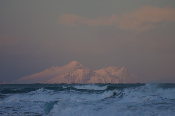Seascape view of pink evening light over snow-covered mountain and breaking waves in sea