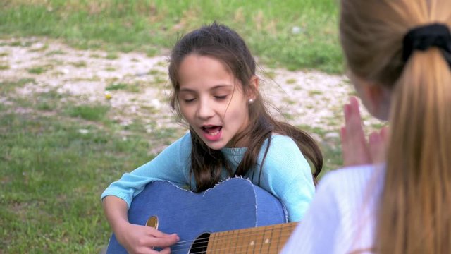 attractive little girl plays acoustic guitar and sings in park while her girlfriend standing listening music performer