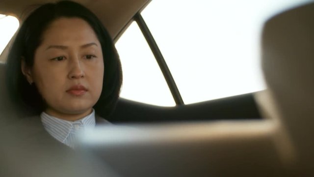 Medium shot of middle-aged Asian woman sitting in backseat of car and working on laptop computer
