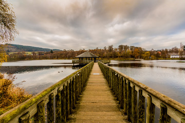 Fototapeta na wymiar Straight wooden bridge towards a gazebo in the middle of Lake Doyards surrounded by autumn greenery, light wind on water surface, cloudy sky covered with clouds at Vielsalm in the Belgian Ardennes