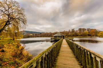 Straight wooden bridge leading to a gazebo in the middle of the Doyards lake surrounded by autumn vegetation, cloudy sky covered with clouds in Vielsalm, the Belgian Ardennes