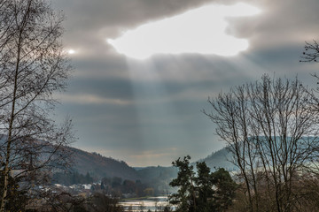 Rays of sunlight between clouds with landscape lake Doyards and village of Vielsalm, mountains covered with trees in background, view from hill between autumnal trees, cloudy day in Belgian Ardennes