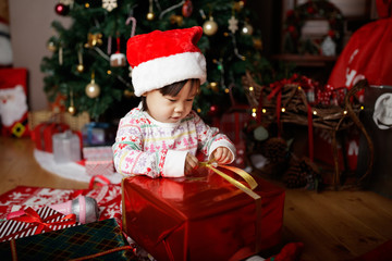 Fototapeta na wymiar toddler baby girl wearing santa claus costume open gift box in front of christmas tree