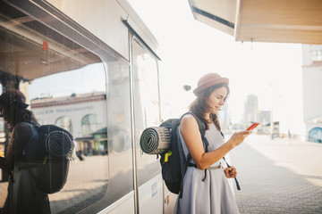 Theme transportation and travel. Portrait young caucasian woman with toothy smile standing train station train background with backpack using technology, smart phone hands in dress and hat summer