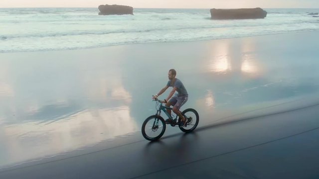 sporty young man riding his bike on the ocean beach on the background of big waves and sunset. Conceptual image of freedom, health and vivacity of spirit. Bali, Indonesia