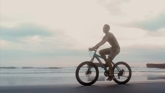 sporty young man riding his bike on the ocean beach on the background of big waves and sunset. Conceptual image of freedom, health and vivacity of spirit. Bali, Indonesia