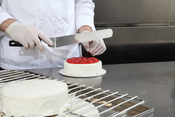 hands pastry chef prepares a cake, cover with icing and decorate with strawberries, works on a stainless steel industrial kitchen work top