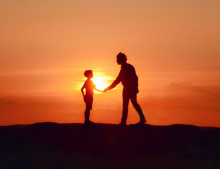 Father and son shake hands and look at each other, idyllic scene in evening landscape with view of setting sun. Dad and boy on fathers day walk together in mountains at beautiful sunset background.