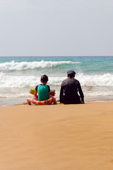 A woman in a Muslim black bathing suit is sitting alone with a girl on the beach.