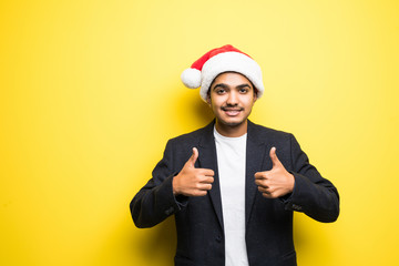 Closeup portrait of young handsome indian man in red santa hat giving thumbs up sign with fingers, isolated on yellow background. Positive emotion facial expressions and symbols