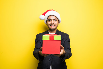 Closeup portrait of happy young handsome man in red santa claus hat holding gift isolated on yellow background.