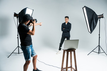 Professional photo shooting at the studio: young man model is smiling and posing the photographer is taking pictures with a digital camera