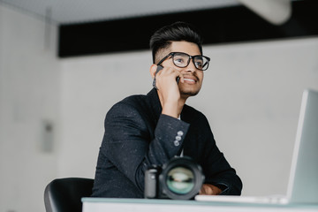 Young photographer working with laptop in office and talking on phone