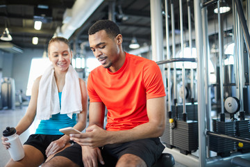 Fototapeta premium Two intercultural athletes in activewear scrolling through previews in smartphone while having break after workout in gym
