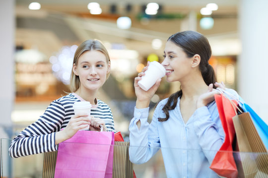 Young women with drinks in plastic glasses deciding where to go next while shopping on xmas sale