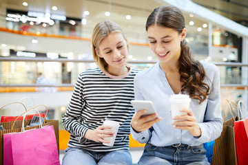 Two girls with drinks searching in online shops and comparing prices with those in offline...