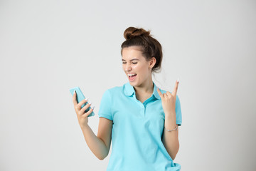 Joyful girl dressed in light blue t-shirt smiles keeps mobile phone in her hand and shows rock sign