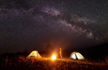 Camping in mountains. Young slim tourist girl standing between two tents watching brightly burning bonfire on deep dark sky with lot of bright sparkling stars background. Tourism and travel concept.