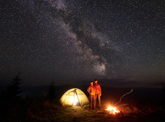 Couple hikers man and woman standing near tent, bonfire under starry sky with Milky way, enjoying quiet night in camping in mountains. Tourism, traveling, outdoor activity and beauty of nature concept