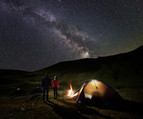 Back view of male and female backpackers holding hands, enjoying milky way on the starry sky at night camping. Beside illuminated tent and campfire is burning. Unusual landscape of mountains and lake