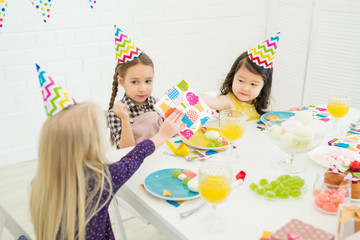 Little friends congratulating birthday girl: cute Asian girl in party hat giving paper napkin to...
