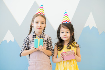 Content cute multi-ethnic little girls in colorful party hats standing against painted wall and holding small gift boxes while looking at camera