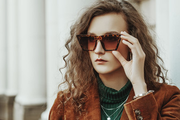 Outdoor close up fashion portrait of young beautiful woman wearing leopard print sunglasses, brown corduroy blazer, green turtleneck, necklace, bracelet, posing in street of european city. Copy space