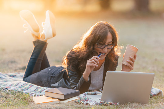 Young woman with eye glasses laying on a the blanket in a park, working with laptop or studying with  notebook and books, and drinking a coffee to go
