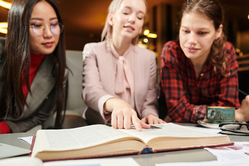 Group of college students sitting at the table and reading a book together or looking for some information in a book