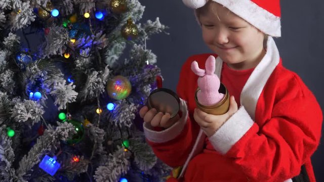 Happy child Dressed in Santa Claus suit, pulls out pink rabbit toy from gift box on background of Christmas tree in garlands. Christmas Eve, New Year