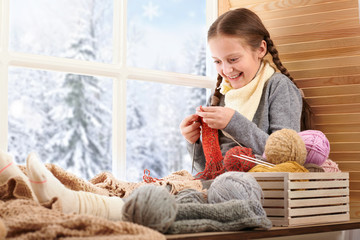 Child girl is sitting on a window sill with wool yarns and knitting. Beautiful view outside the...