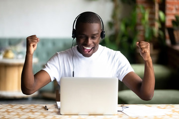 Excited smiling African American man in headphones celebrating success raise hands, online win...
