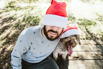 .Young man with his dog friend, both wearing santa claus caps celebrating Christmas on a sunny December day. Lifestyle