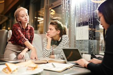 Group of female friends gathered in cafe and doing homework together, using literature books and laptop computer