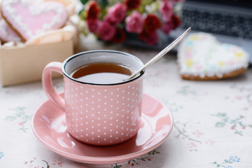 Hot tea in a pink cup on a pink saucer against the background of a bouquet of flowers, heart-shape gingerbreads and a laptop keyboard in Valentin's day.