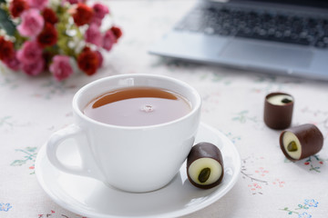 Hot tea in a white cup next to chocolate candy on a white saucer against the background of a bouquet of flowers and a laptop keyboard and candy.