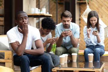 Bored African American man sitting with people with phone dependence, yawning, friends using mobile...