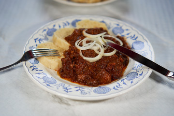 Slow cooked czech or hungarian beef goulash stew served with traditional czech dumplings and choped fresh onion on the rustic plate with stainless steel fork and knive.