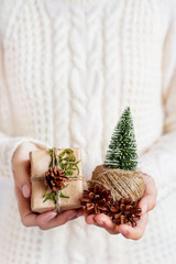 Woman in white knitted sweater holding a present packed in craft paper with pine cone. DIY Christmas or New Year, St. Valentine Day gift.