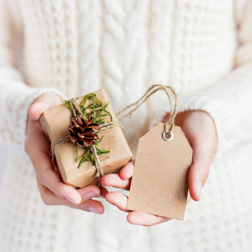 Woman in white knitted sweater is holding a present packed in craft paper with pine cona and tag. DIY Christmas or New Year, St. Valentine Day gift.