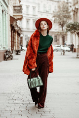 Outdoor full body fashion portrait of young beautiful woman wearing trendy oversized orange faux fur coat, hat, green sweater, corduroy trousers, holding stylish snakeskin bag, posing in street 