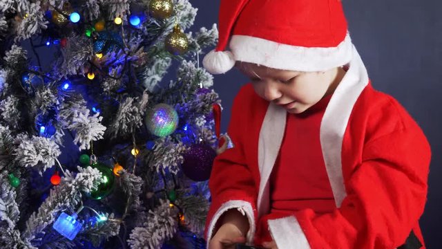 Happy child Dressed in Santa Claus suit, pulls out pink rabbit toy from gift box on background of Christmas tree in garlands. Christmas Eve, New Year