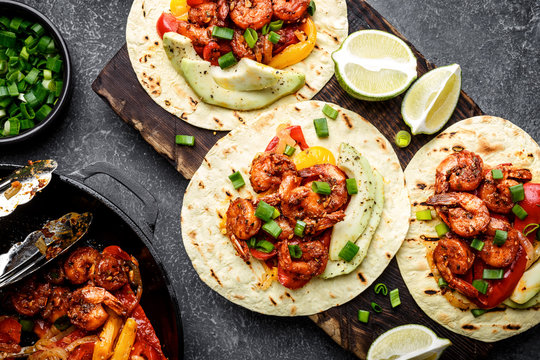 Fajitas in tortillas with fried shrimps, bell peppers and onion served up with avocado and green onions on wooden cutting board, top view, flat lay