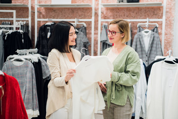 Beautiful girls are choosing clothes, talking and smiling while doing shopping in boutique