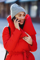 Happy fashion woman using a smart phone and talking wearing red coat like down jacket and grey knitted hat in the street in winter - Image. Winter holiday cocept and communication