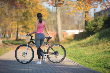 Obraz premium Young beautiful girl in sportswear stands next to a bicycle on the street in the park in autumn.