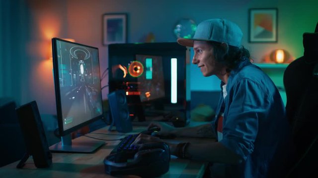 Joyful Gamer Playing First-Person Shooter Online Video Game on His Personal Computer. Room and PC have Colorful Neon Led Lights. Young Man is Wearing a Cap. Cozy Evening at Home.